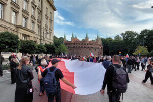 Uczestnicy marszu niosą w poziomie wielką flagę Polski. W tle widać Barbakan.